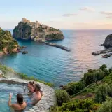 Coppie rilassate in una sorgente di nitrodi gratis con vista su un castello e un borgo sul mare.