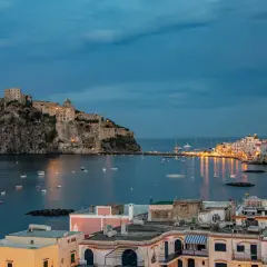 Castello Aragonese domina il mare blu, con barche che ondeggiano e luci che illuminano le pittoresche calette di Ischia al crepuscolo.