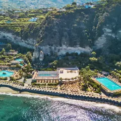 Vista aerea di un resort con piscine e lettini sulla spiaggia, incastonato tra il mare e una scogliera verdeggiante. Un paradiso di terme a Forio.