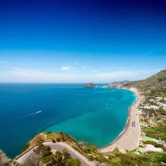 Vista panoramica delle spiagge libere a Ischia, con il mare turchese che lambisce la costa e le case arroccate sulle colline verdi.