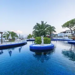 Piscina azzurra con isolotto centrale e statua, lettini e palme intorno. Un'oasi di relax all'Hotel Apollon.