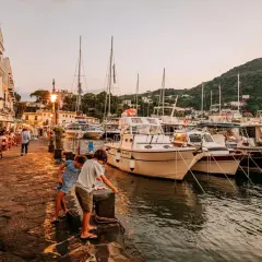 Bambini giocano sul molo della riva destra Ischia, con ristoranti affollati e barche ormeggiate al tramonto.
