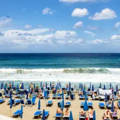 Ischia ad agosto: una spiaggia affollata con lettini blu, ombrelloni e bagnanti che si godono il mare cristallino sotto un cielo azzurro con nuvole bianche.