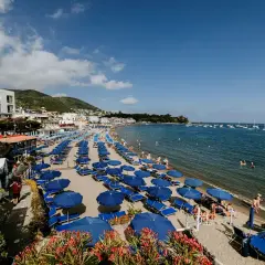 Spiaggia affollata a Ischia Lido con file di ombrelloni blu e lettini, bagnanti in acqua e barche all'orizzonte.