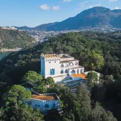Villa La Colombaia a Ischia, un bianco maniero con torrette e finestre blu, immerso nel verde lussureggiante con vista sul mare e sulle montagne.