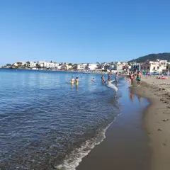 Persone si godono una giornata di sole sulla spiaggia di San Pietro, con il mare azzurro e case colorate sullo sfondo.