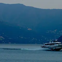 Aliscafo in navigazione su un lago calmo, con montagne boscose e case illuminate sullo sfondo.