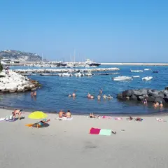 Spiaggia della Marina di Ischia: bagnanti si godono il sole e il mare, con barche ormeggiate nel porto sullo sfondo.