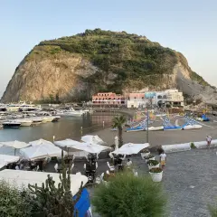 Il porto turistico di Ischia, con yacht ormeggiati, una spiaggia con lettini e ombrelloni, e case colorate ai piedi di una collina rocciosa.