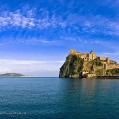 Il **palazzo reale** di Ischia, arroccato su uno scoglio, domina il mare blu sotto un cielo terso.