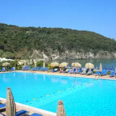 Piscina azzurra con lettini e ombrelloni, vista sul mare e collina verdeggiante. Un angolo di paradiso, forse Negombo o un luogo ispirato a Poseidon.