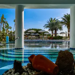 Piscina azzurra con palme e lettini, un'atmosfera da **parco termale romantica** con un'antica anfora come decorazione.