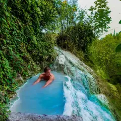 Un uomo si rilassa nelle acque azzurre di una cascata termale naturale a Olmitello, Ischia, circondato da lussureggiante vegetazione.