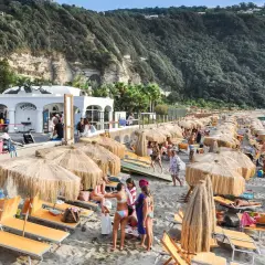 Spiaggia di Citara affollata, con ombrelloni di paglia, lettini arancioni e gente che si gode il mare. Per arrivare, segui le indicazioni per Forio.