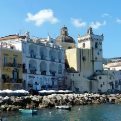 Colorate case affacciate sul mare, un porto con barche e rocce. Per sapere come arrivare ad Ischia in treno, si può prendere un treno per Napoli e poi un aliscafo.