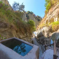 Piscina naturale scavata nella roccia, parte delle storiche terme di Cavascura, circondata da vegetazione e pareti rocciose.