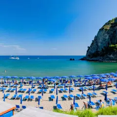 Spiaggia affollata con ombrelloni blu e lettini sulla riviera di San Montano. Mare azzurro e scogliera verde sullo sfondo.