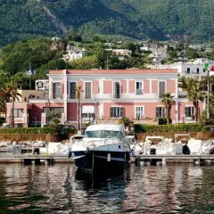 Il **hotel Svizzera Ischia** si affaccia sul porto con barche ormeggiate. Sullo sfondo, case arroccate su una collina verdeggiante.
