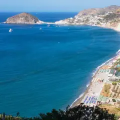 Vista panoramica della spiaggia dei Maronti con lidi attrezzati, ombrelloni colorati e il mare blu intenso. In lontananza, un isolotto e la costa frastagliata.