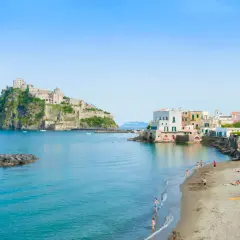 Lidi di Ischia Porto con il Castello Aragonese sullo sfondo. Gente in spiaggia e mare azzurro sotto un cielo limpido.