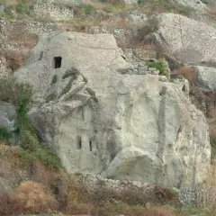 Antiche case in pietra scavate nella roccia e costruite con muretti a secco, immerse in un paesaggio collinare brullo.