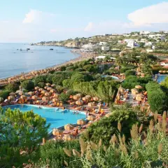 Vista panoramica di un resort a Ischia con piscine, lettini, ombrelloni e la spiaggia che si estende verso il mare.