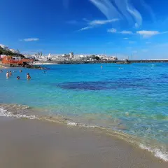 Persone nuotano nelle acque cristalline della spiaggia della Chiaia, con un borgo bianco sullo sfondo e un cielo azzurro con nuvole bianche.