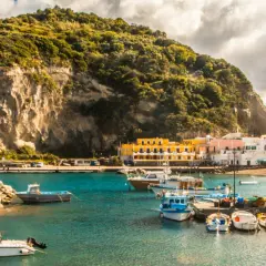 Un pittoresco ormeggio a Ischia, con barche colorate ormeggiate davanti a case bianche e gialle, sotto una collina verdeggiante.