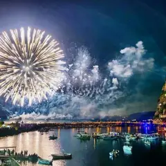 Fuochi d'artificio spettacolari illuminano il cielo notturno sopra un porto pieno di barche, con un castello arroccato su una scogliera. Un'immagine che ricorda le celebrazioni di Mario Mazzella.