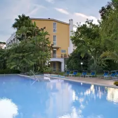 Piscina azzurra e lettini blu in un rigoglioso giardino tropicale, con un edificio giallo sullo sfondo, a San Giovanni, Ischia.
