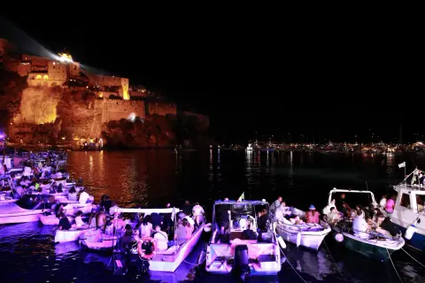 Una magica festa a Ischia: barche illuminate e gente festante con il castello illuminato sullo sfondo.