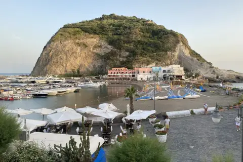 Il porto turistico di Ischia, con yacht ormeggiati, una spiaggia con lettini e ombrelloni, e case colorate ai piedi di una collina rocciosa.