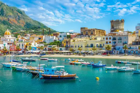 Barche a vela e a motore ormeggiate nel blu intenso del mare, con case colorate e una torre antica a fare da sfondo a Ischia Porto.