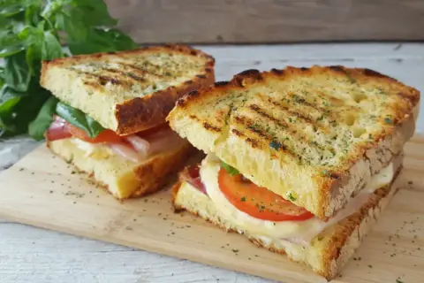 Due panini grigliati, una zingara ischitana, con pomodoro, mozzarella filante e prosciutto, su tagliere di legno.