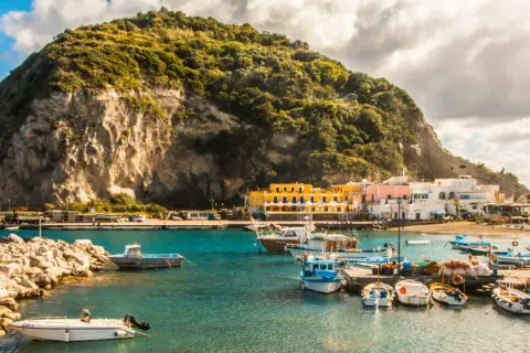Un pittoresco ormeggio a Ischia, con barche colorate ormeggiate davanti a case bianche e gialle, sotto una collina verdeggiante.