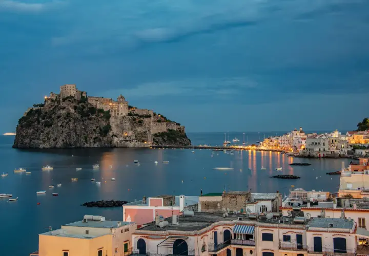 Castello Aragonese domina il mare blu, con barche che ondeggiano e luci che illuminano le pittoresche calette di Ischia al crepuscolo.