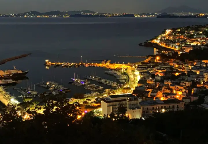 Casamicciola, un gioiello illuminato di notte, con un porto pieno di barche e luci che si riflettono sul mare calmo.