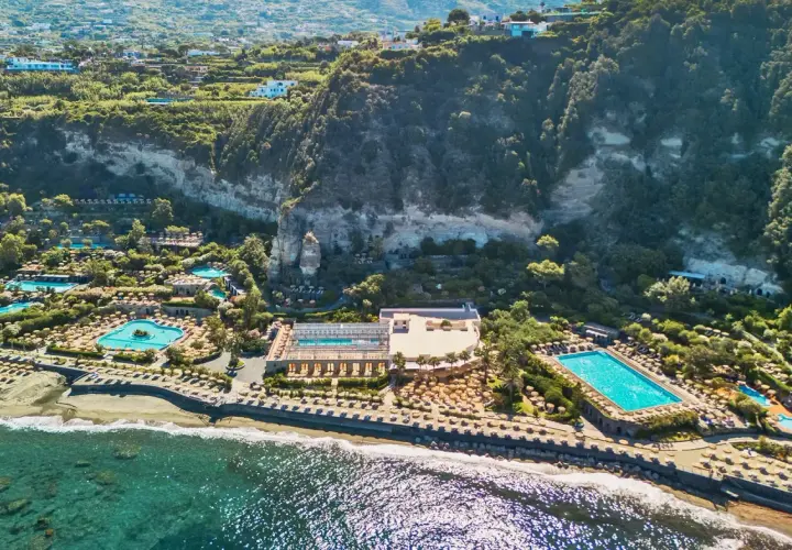 Vista aerea di un resort con piscine e lettini sulla spiaggia, incastonato tra il mare e una scogliera verdeggiante. Un paradiso di terme a Forio.