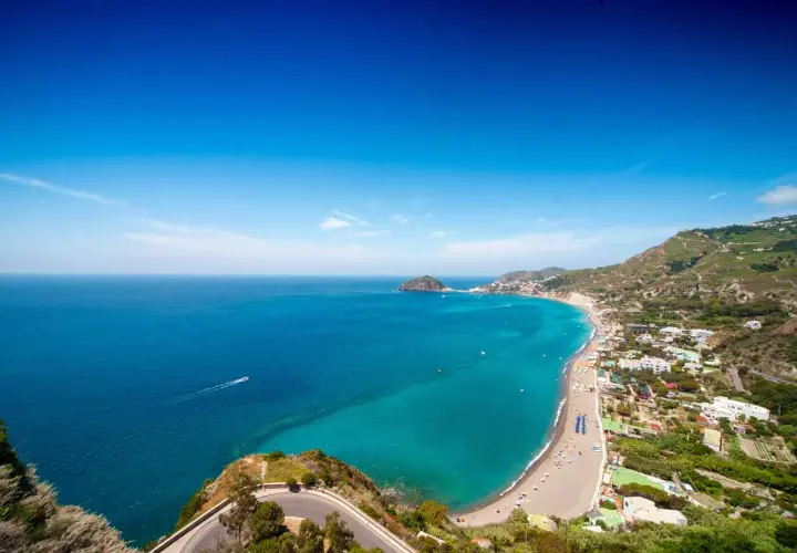 Vista panoramica delle spiagge libere a Ischia, con il mare turchese che lambisce la costa e le case arroccate sulle colline verdi.