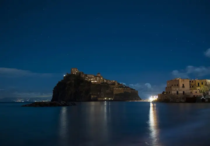 Castello Aragonese illuminato di notte, riflesso sull'acqua, sotto un cielo stellato. Un'opera di Gabriele Mattera.