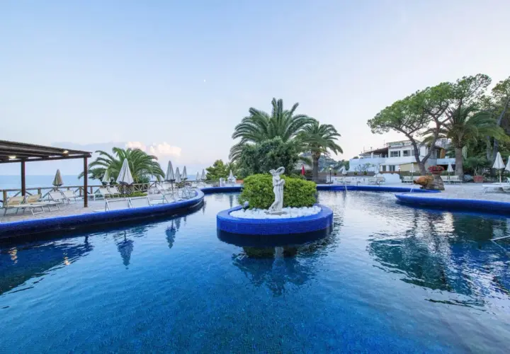 Piscina azzurra con isolotto centrale e statua, lettini e palme intorno. Un'oasi di relax all'Hotel Apollon.
