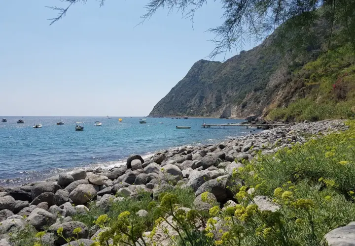La baia della Scarrupata, con le sue rocce levigate e il mare azzurro, accoglie piccole imbarcazioni. Un pontile si protende nell'acqua.