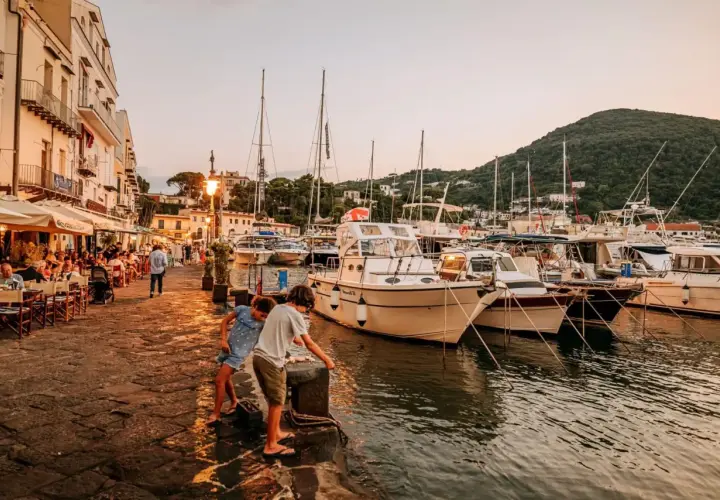 Bambini giocano sul molo della riva destra Ischia, con ristoranti affollati e barche ormeggiate al tramonto.