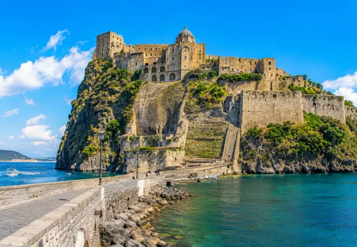 The Aragonese Castle on a rocky island, connected by a stone bridge, with boats in the turquoise sea.
