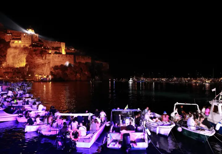 Una magica festa a Ischia: barche illuminate e gente festante con il castello illuminato sullo sfondo.