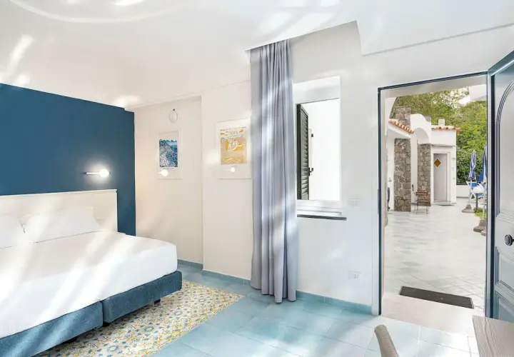 Camera luminosa con letto matrimoniale e vista su un cortile con ombrelloni blu. Un invito a soggiornare all'hotel oasi Ischia.