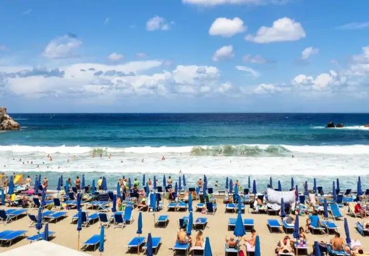 Ischia ad agosto: una spiaggia affollata con lettini blu, ombrelloni e bagnanti che si godono il mare cristallino sotto un cielo azzurro con nuvole bianche.