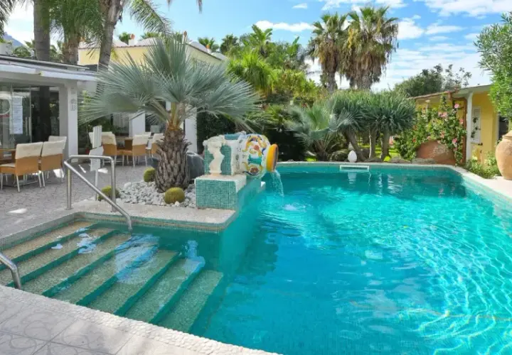 Piscina cristallina con cascata decorativa, lettini prendisole e area ristoro esterna. Un angolo di relax che ricorda le terme comunali di Ischia.