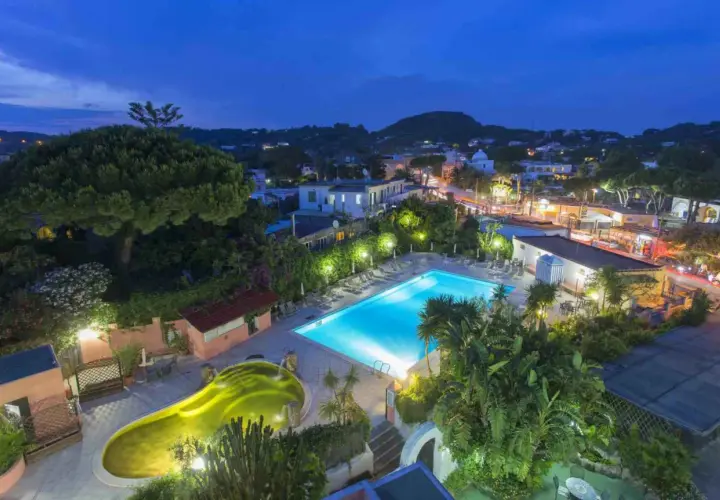 Piscina illuminata e vasca idromassaggio a forma di fagiolo al Hotel Eden Ischia, con vista sul paese di sera.
