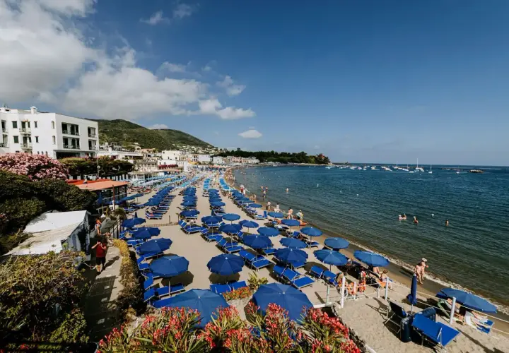 Spiaggia affollata a Ischia Lido con file di ombrelloni blu e lettini, bagnanti in acqua e barche all'orizzonte.